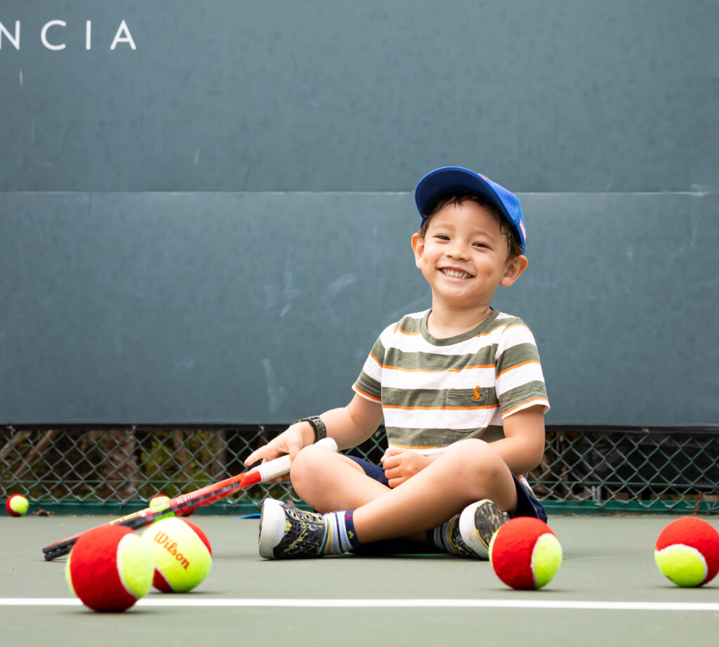 little boy sitting on a tennis court surrounded by tennis balls