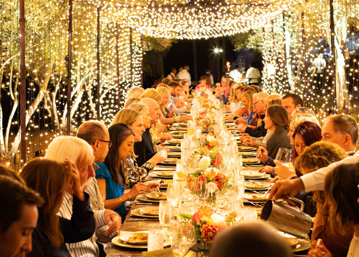 a long dining table full of people with string lights overhead