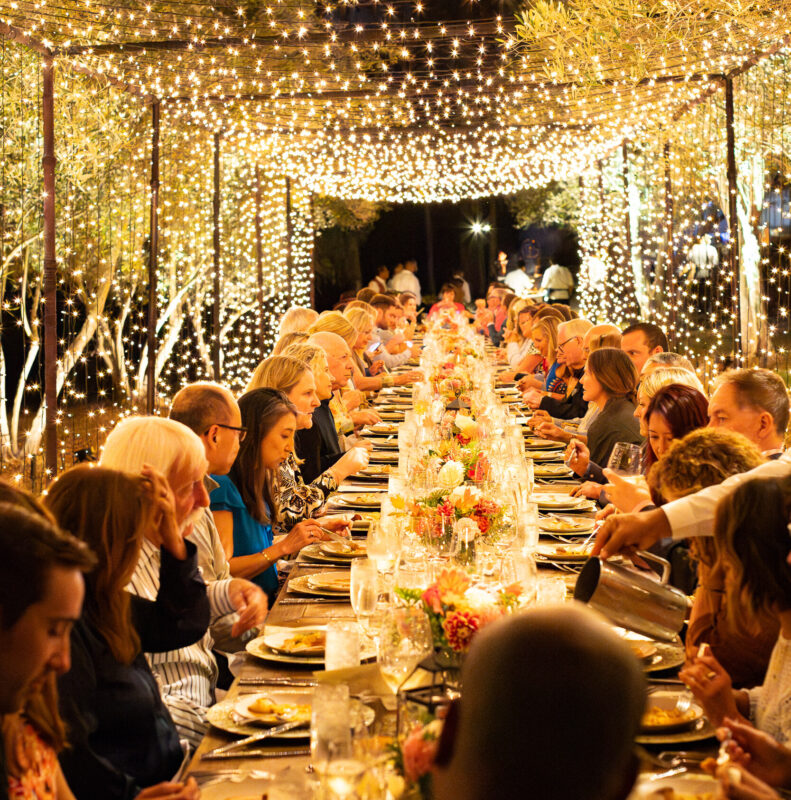 a long dining table full of people with string lights overhead in the culinary garden